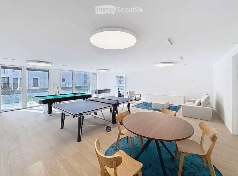 Bright, modern room with large windows, featuring a sofa, a round wooden table with chairs, a ping pong table, and a pool table.