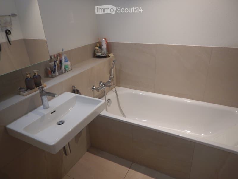 Modern bathroom with a white sink, bathtub, and mirror. The sink has a chrome faucet, and the bathtub features a handheld showerhead. A toothbrush holder and small bottles are placed on the windowsill.