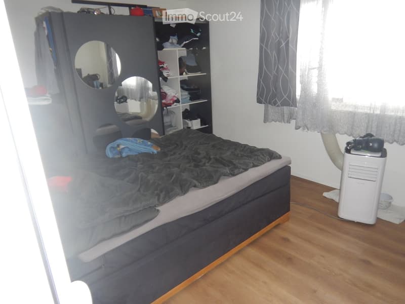 Bedroom with a wooden floor, a bed with dark bedding, a wardrobe with mirrors, a shelf, and an air conditioner unit near the window.