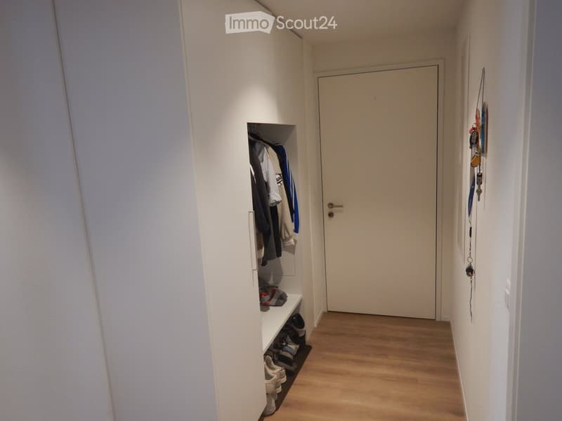 A narrow hallway with light wooden flooring, white walls, a built-in shoe rack, and a closed door at the end.