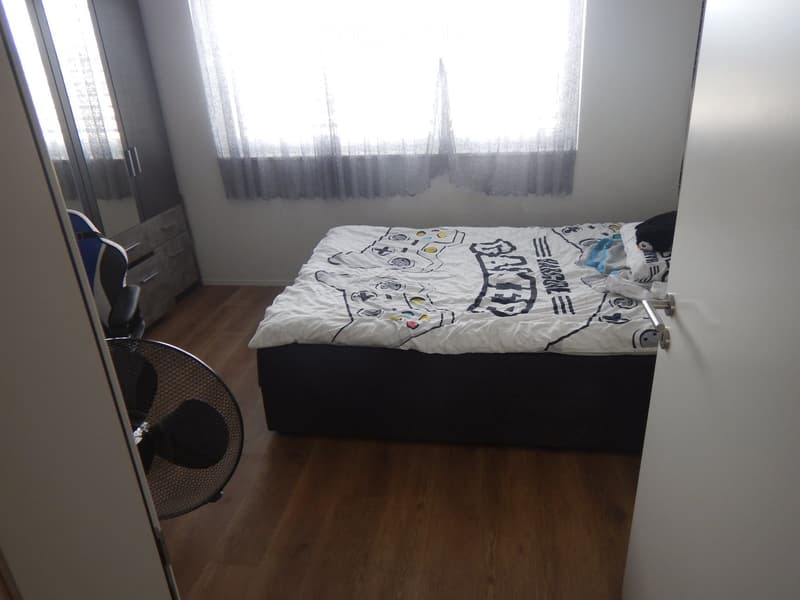A small bedroom with wooden flooring, a bed with a colorful duvet, a fan, and a chair near the window.