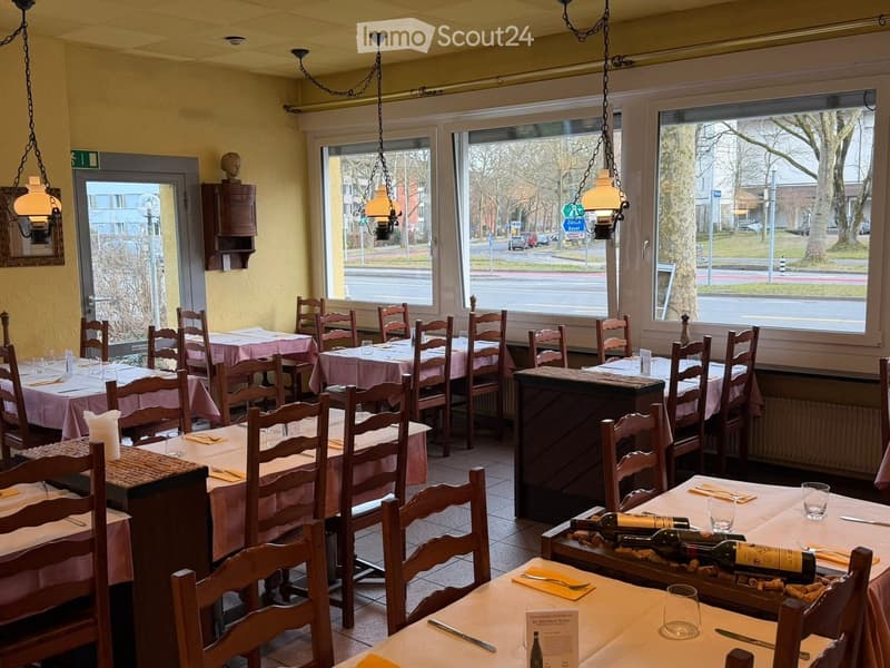 Restaurant interior with multiple tables and chairs, each set with wine bottles, menus, and cutlery. Large windows provide a view of the street outside.