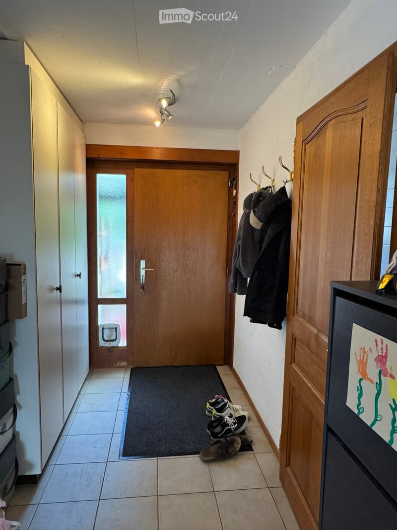 Hallway with tiled floor, wooden doors, coat hooks with jackets, shoes on the floor, and a small pet door with a camera.