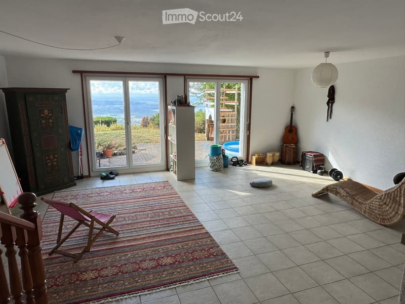 Bright living space with large glass doors opening to a scenic view, featuring a colorful rug, wooden chair, guitar, and various decorative items.