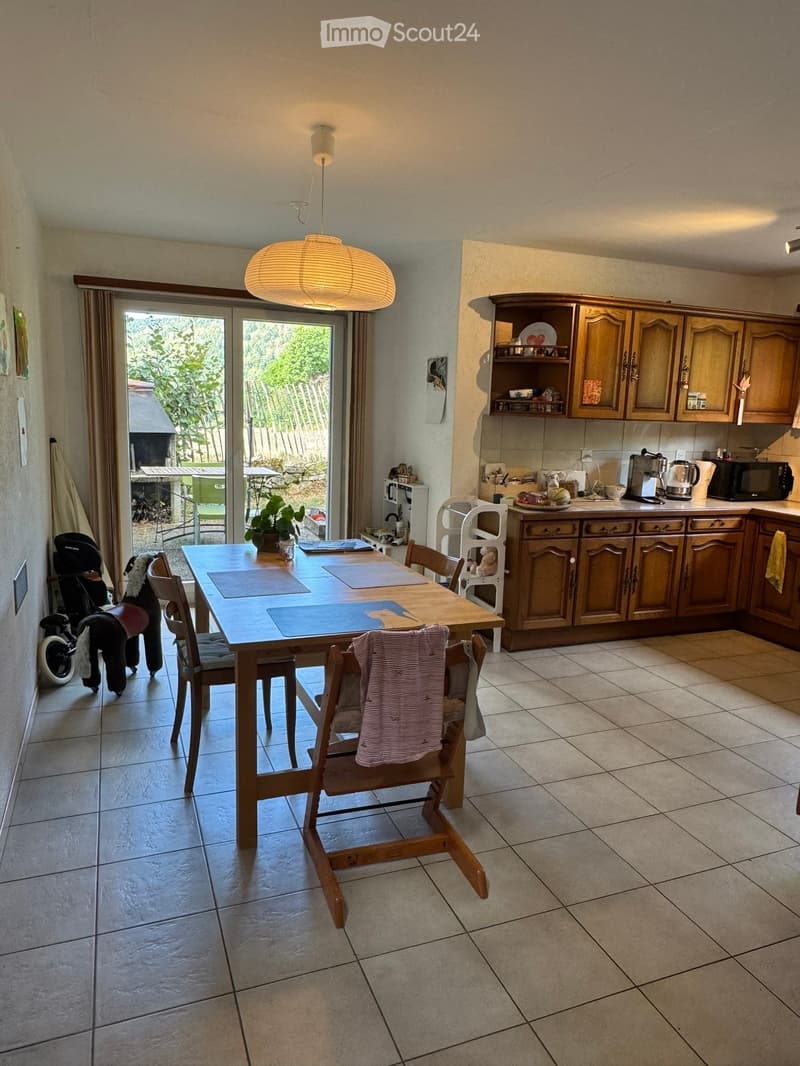 Dining area with a wooden table, chairs, and a kitchen area with cabinets, microwave, and coffee maker. A large window with a view of the garden is visible.