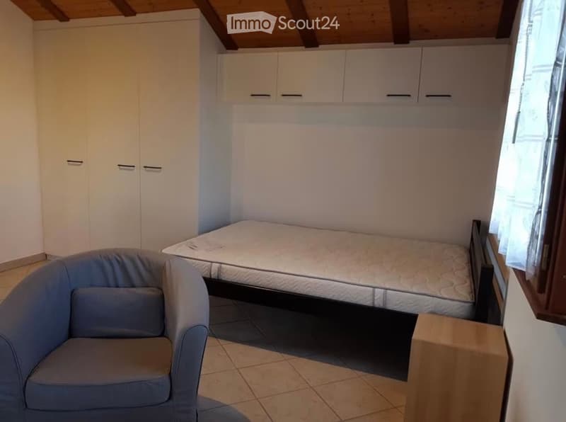 Cozy bedroom with a single bed, a grey armchair, and built-in white wardrobes. The room features a slanted wooden ceiling and tiled flooring.