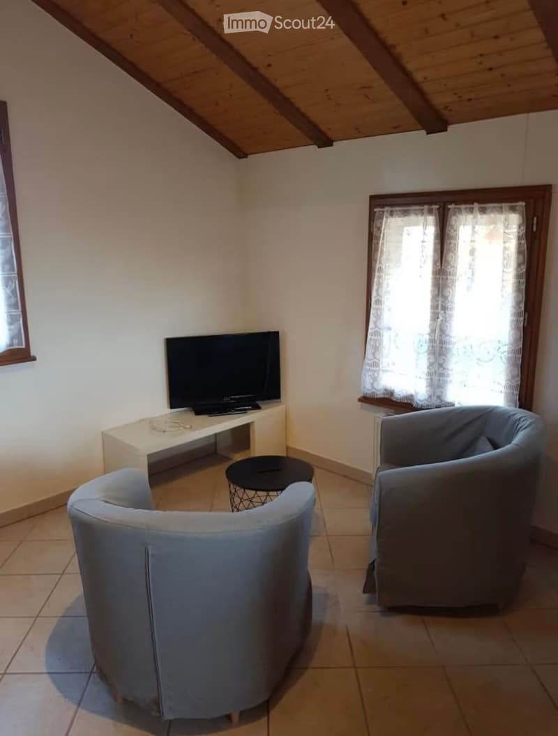 Living room with two blue armchairs, a small black table, a white TV stand, and a flat-screen TV. The room has wooden beams on the ceiling and large windows with white curtains.
