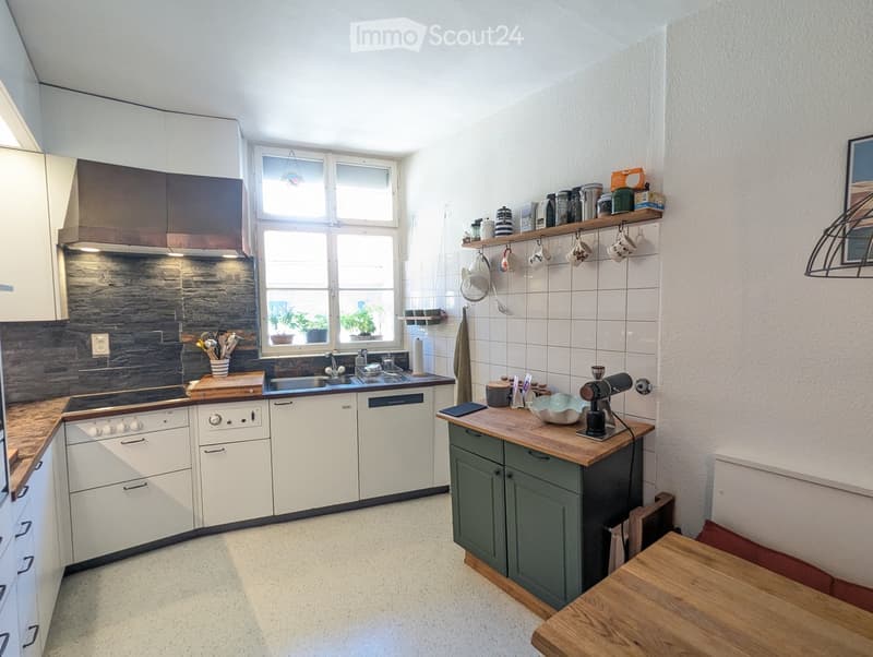 Modern kitchen with white cabinets, a central island, and a window overlooking a small garden. Includes a stove, dishwasher, sink, and various kitchen utensils.