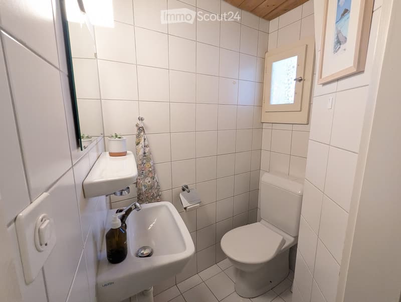 Small bathroom with white tiles, a sink, toilet, mirror, and a small window above the toilet.