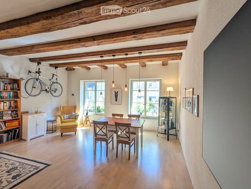 Bright, open room with wooden beams, hardwood floor, large windows, dining table with chairs, yellow armchair, bookshelf, bicycle mounted on wall, glass display cabinet, and table lamp.