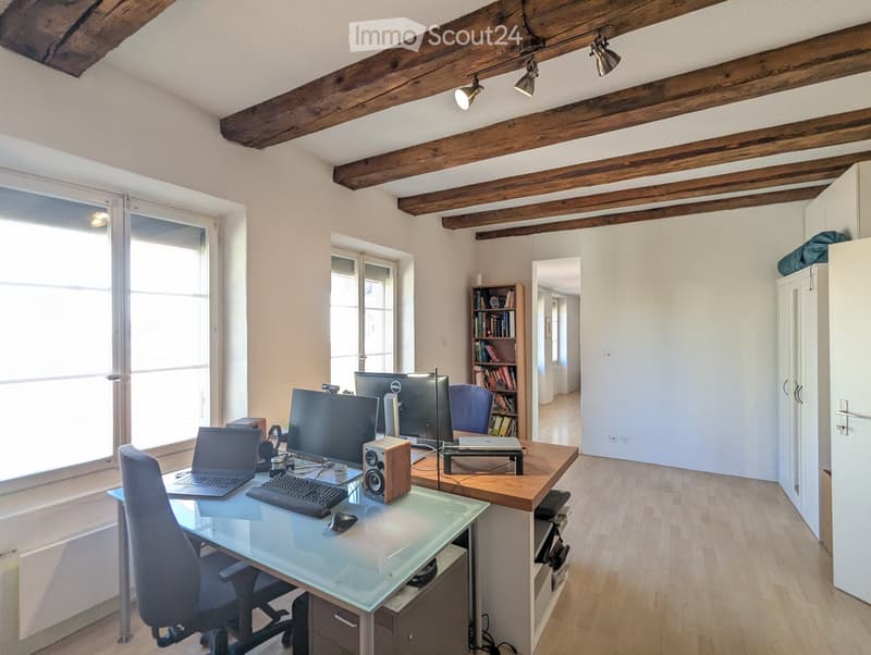 A modern office space with a glass desk, laptop, keyboard, mouse, and a bookshelf filled with books. The room features wooden beams on the ceiling and large windows allowing natural light to fill the space.