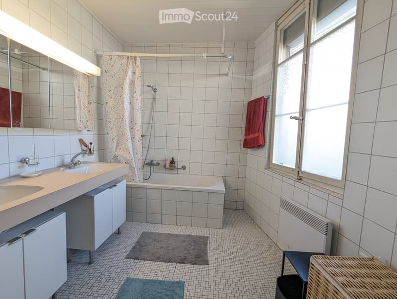 Bathroom with white tiled walls and floor, double sink, bathtub, mirror, towel, and window with blinds.