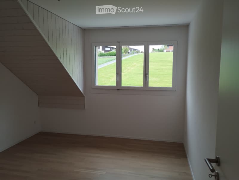 Empty room with wooden flooring, white walls, and large windows overlooking a green lawn.