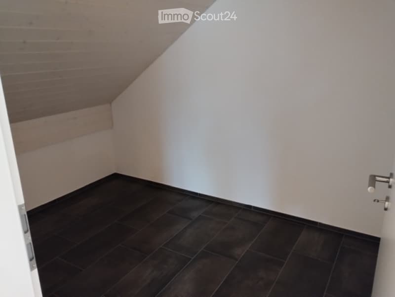 Completely empty room with dark tiled floor and white walls, featuring a slanted wooden ceiling and a door frame visible on the left side.