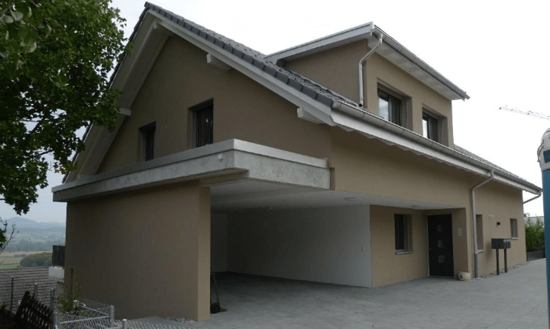 Two-story house with beige exterior, garage, and multiple windows. The house features a modern design with a sloped roof and a raised ground floor.