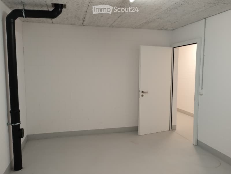 Empty room with white brick walls, concrete floor, and a single white door leading to another space. A large black pipe is mounted on the left wall.