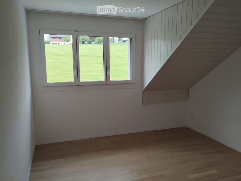 Empty room with wooden flooring, white walls, and large windows overlooking a green lawn.