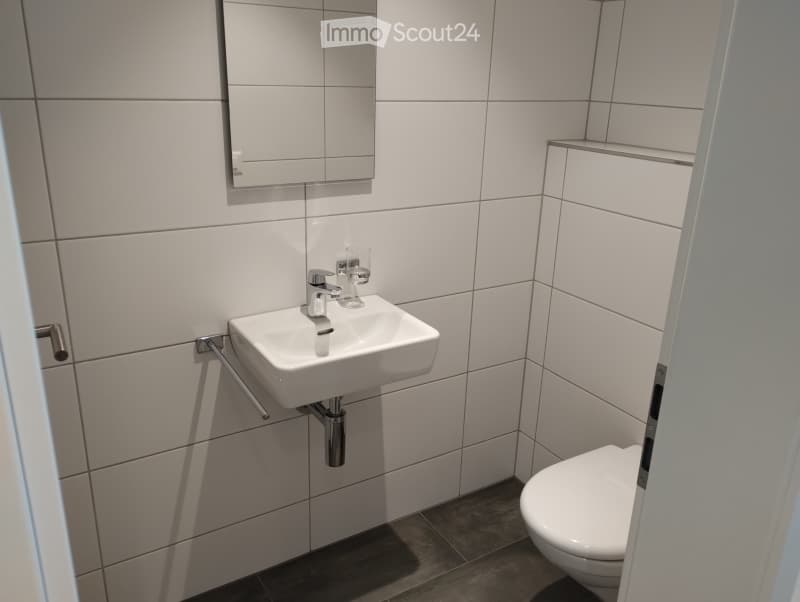 Modern bathroom with white tiles, a white sink, a mirror, and a toilet. The sink has a chrome faucet and a towel rack is mounted on the wall.