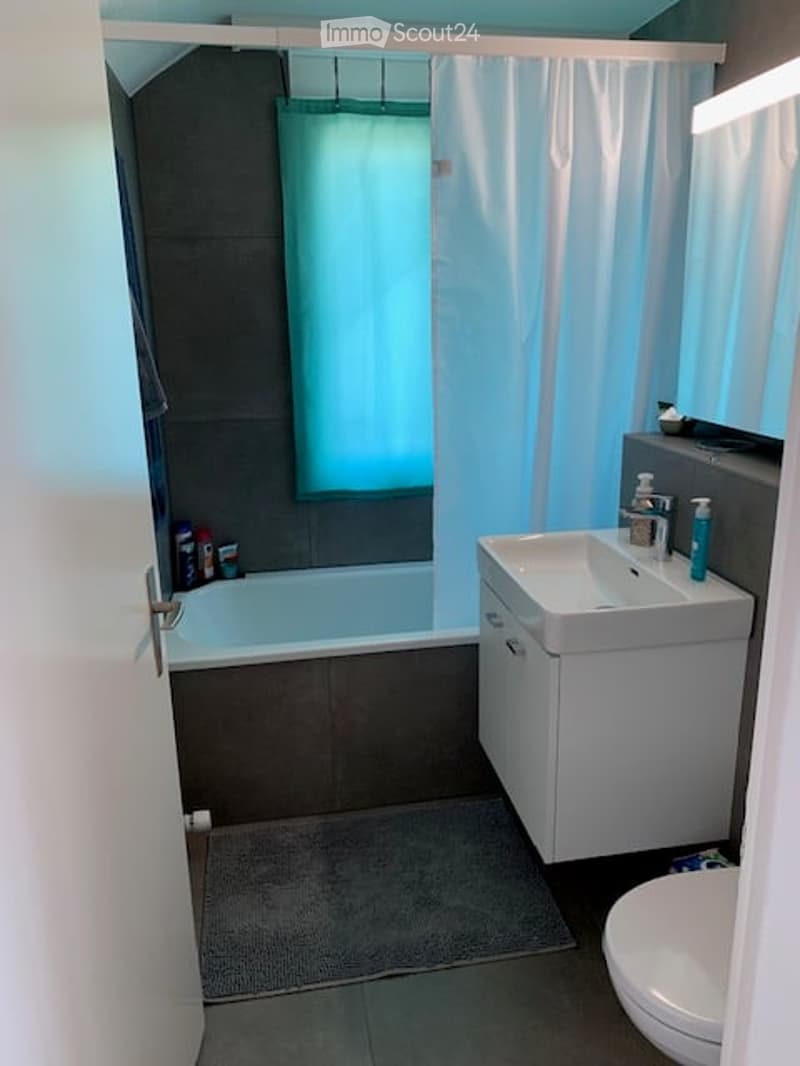 Modern bathroom with a bathtub, sink, toilet, and a large mirror above the sink. The space features a grey color scheme with a white cabinet and a blue shower curtain.