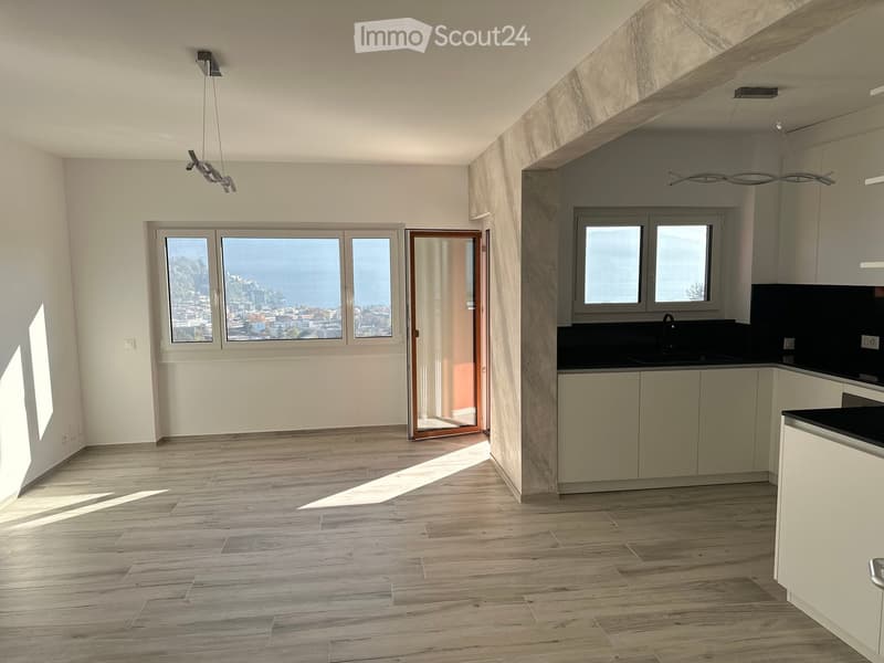 Bright, open-plan living and dining area with wooden flooring, large windows offering a scenic view, and a modern kitchen area with light-colored cabinets and a black countertop.