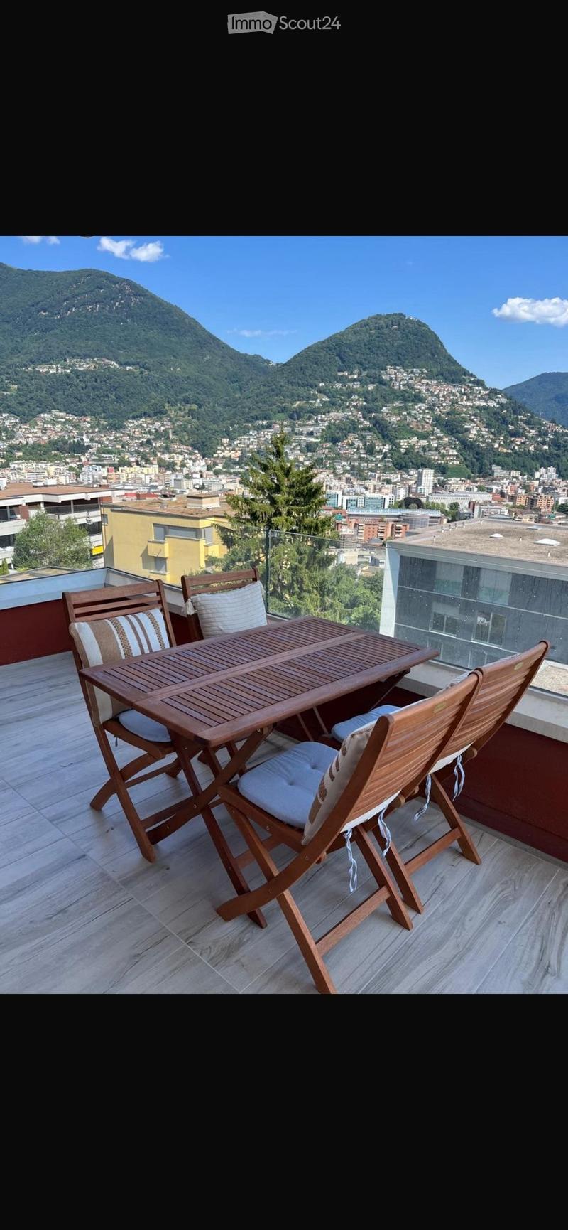 Balcony with wooden table and chairs, overlooking a scenic cityscape and mountains.