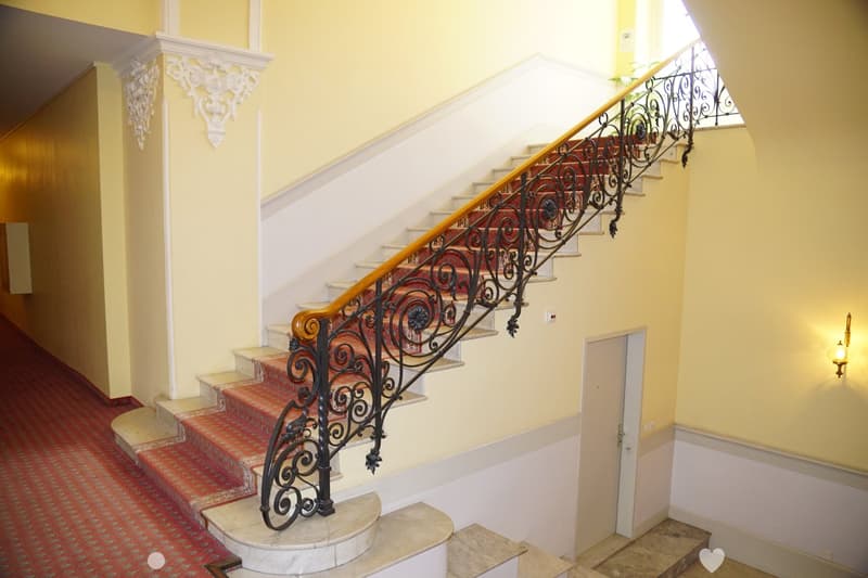 Elegant staircase with ornate wrought iron railing, red patterned carpet, and light yellow walls.