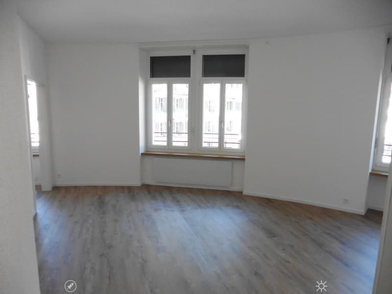 Empty room with wooden flooring, white walls, and large windows with a view of a building outside.