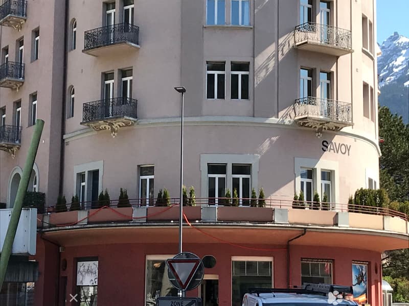 Multi-story building with balconies, large windows, and decorative railings. The facade is light-colored with the word "SAVOY" visible. The lower level has a storefront with signage and a vehicle parked in front.