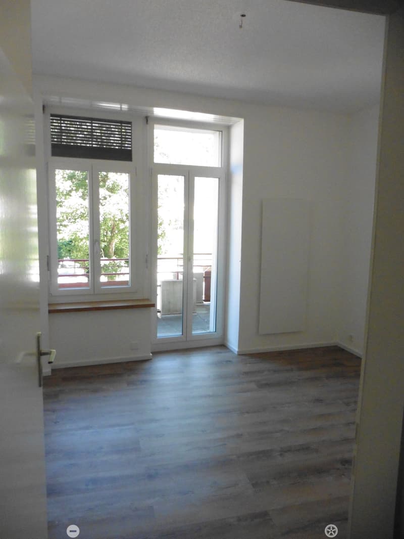 Empty room with wooden flooring, white walls, large windows, and a sliding glass door leading to a balcony.