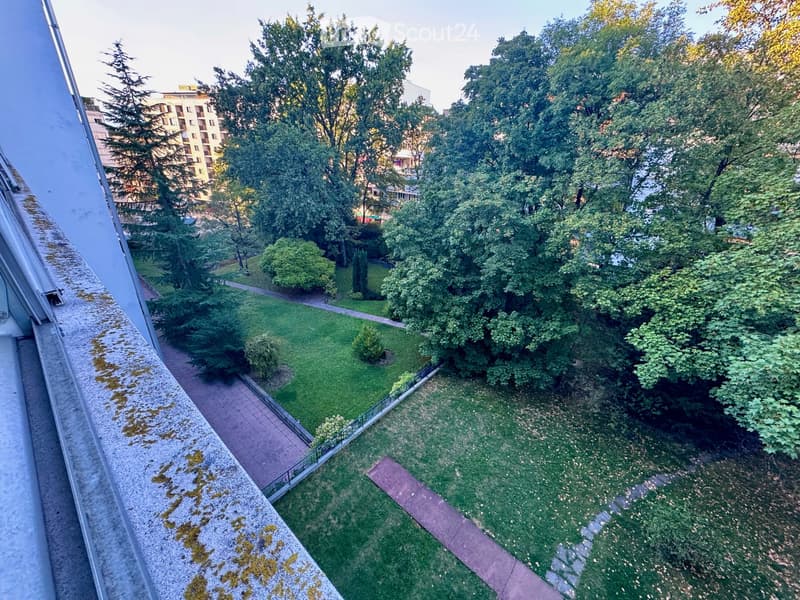 Balcony overlooking a green park area with trees, pathways, and a building in the background.