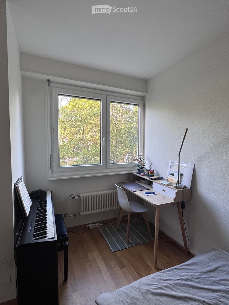 Cozy bedroom with a bed, desk, chair, piano, and a window view of greenery.