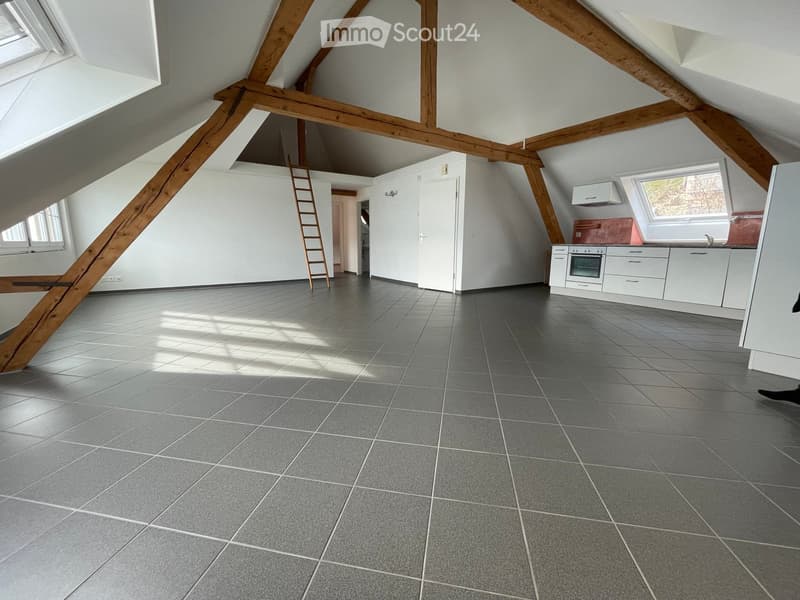 Bright attic space with exposed wooden beams, grey tiled floor, white kitchen area with cabinets, oven, and sink, and a wooden ladder leading to an upper level.