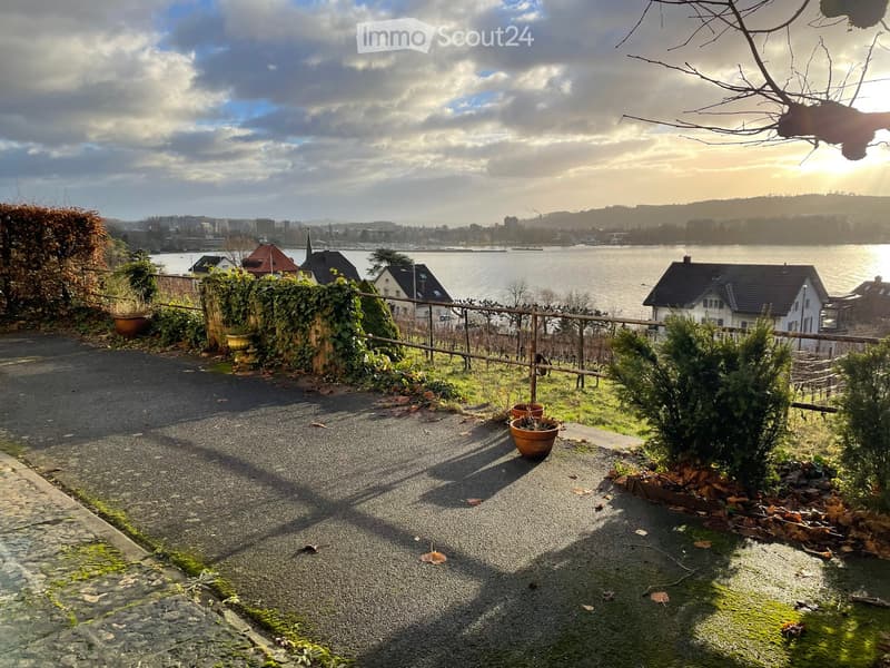 Garden path with potted plants, overlooking a waterfront with houses and a distant cityscape.