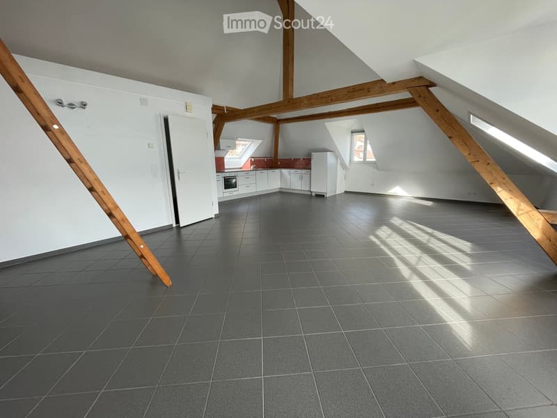 An empty attic space with exposed wooden beams, grey tiled floor, and a small kitchen area visible in the background.