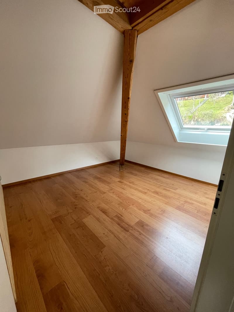 Empty attic room with wooden floor, slanted wooden ceiling, and a skylight window showing greenery outside.