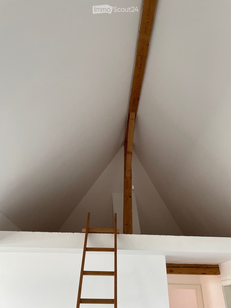 Empty attic space with exposed wooden beams, white walls, and a wooden ladder leading up to the attic floor.