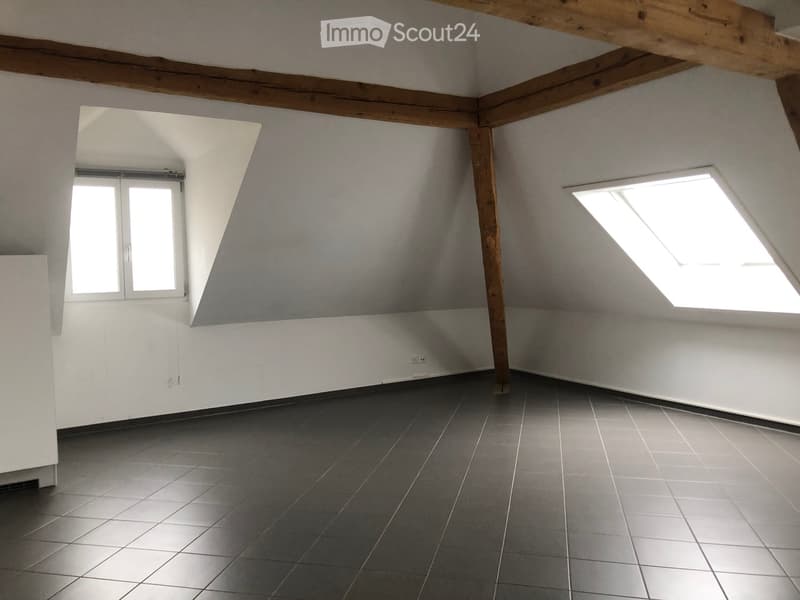 Empty attic room with exposed wooden beams, grey tiled floor, and two windows including a skylight.