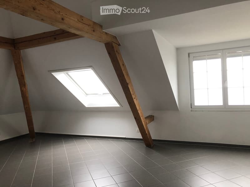 Empty attic room with exposed wooden beams, grey tiled floor, and skylight window providing natural light.