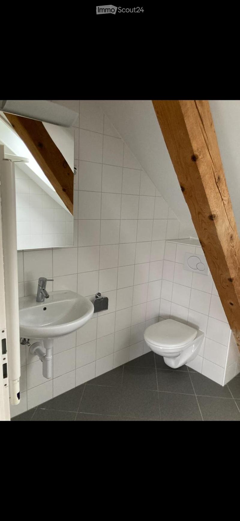 Modern bathroom with white tiles, a sink, a toilet, and a dark grey floor. The room features exposed wooden beams and a small window.