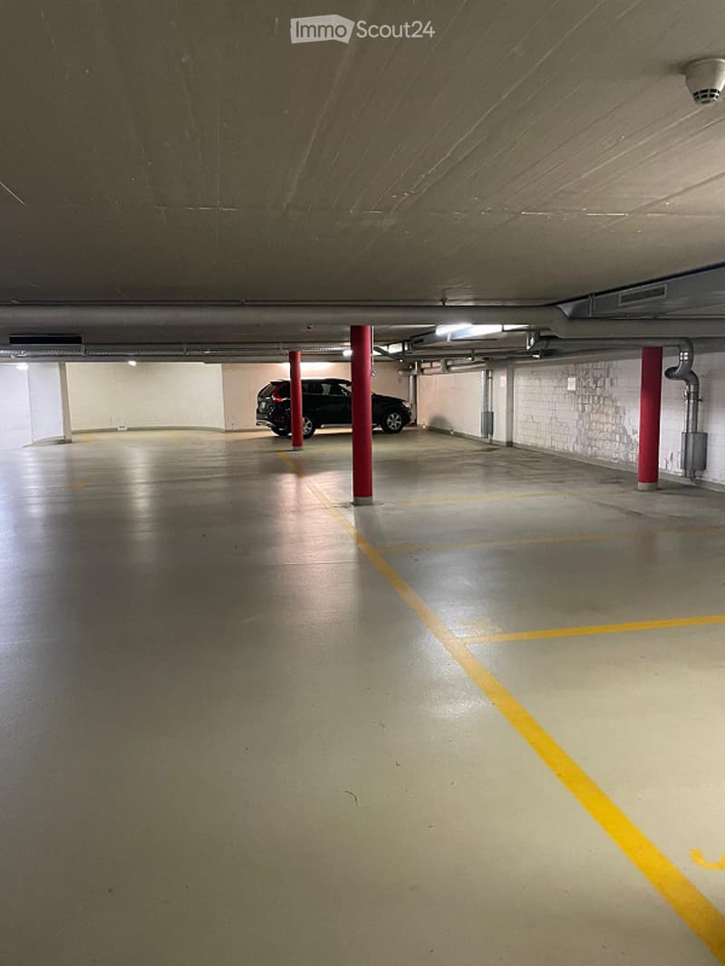 Underground parking garage with concrete floors, red support pillars, and a parked car in the background.