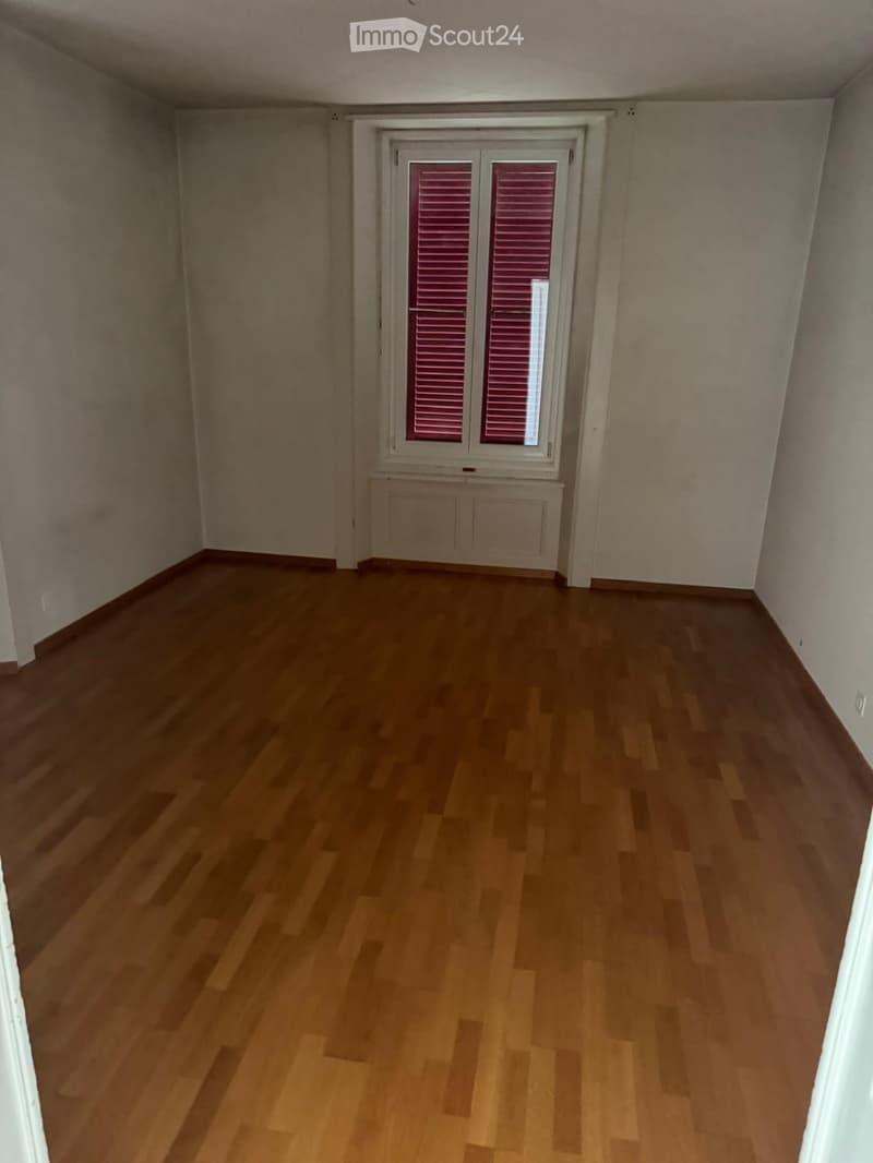 Empty room with wooden flooring, white walls, and a window with pink shutters.