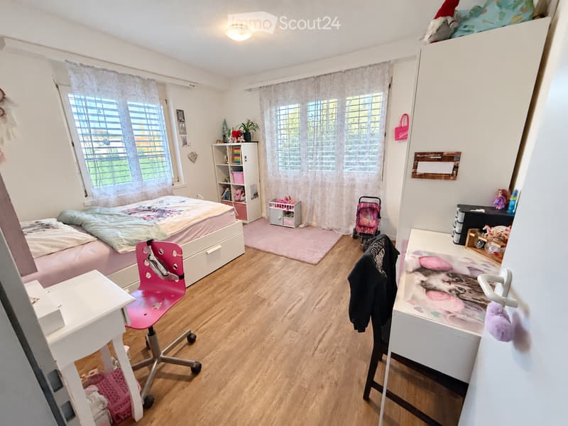 A well-lit bedroom with a bed, a pink chair, a desk, and a shelf. The room features wooden flooring, large windows with curtains, and various decorative items including toys and a plant.
