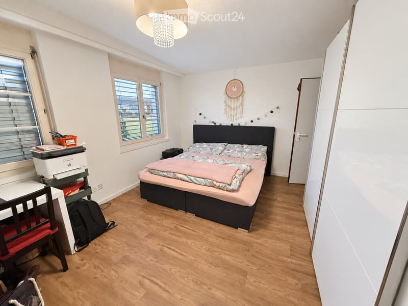 Cozy bedroom with wooden flooring, a double bed with a pink blanket, a desk with a chair, a printer, and a backpack on the floor.