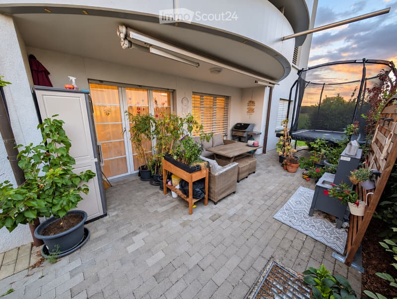 Balcony with tiled floor, outdoor furniture including a table, chairs, and potted plants. Features a trampoline, barbecue, and a view of the evening sky.