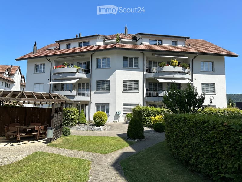 Two-story white house with red-tiled roof, multiple windows, balconies with flower boxes, and a well-maintained garden with a wooden pergola and seating area.