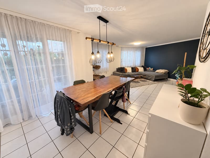 Modern open-plan living and dining area with a wooden dining table, chairs, sofa, ottoman, side tables, pendant lights, and plants.