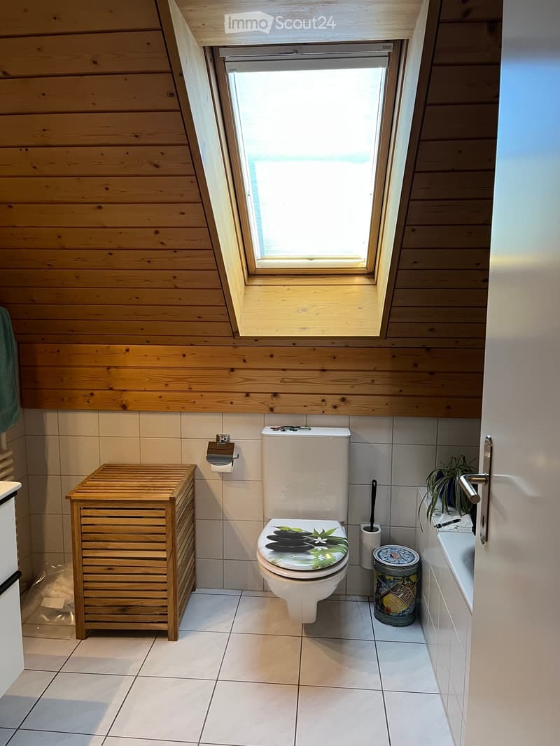 Bathroom with wooden walls, a toilet with a decorative seat cover, a wooden cabinet, a toilet paper holder, a toilet brush, a small plant on the windowsill, and a bathtub with a chrome faucet.