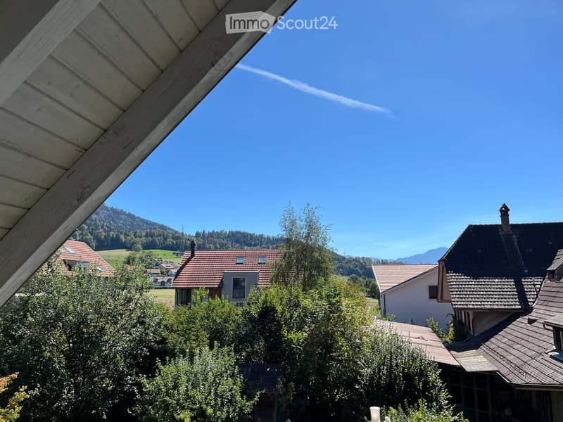 View from a balcony or terrace showing a rural landscape with houses, trees, and distant mountains under a clear blue sky.