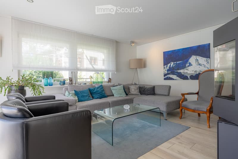 Modern living room with grey sectional sofa, glass coffee table, wooden chair, floor lamp, and large painting on the wall. Large windows with roller blinds and potted plants on the windowsill.