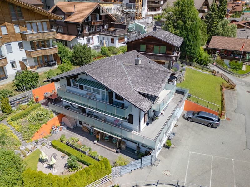 Aerial view of a multi-story house with a grey roof, white walls, and a balcony. The property includes a driveway with a parked car, a well-maintained garden, and outdoor seating areas.